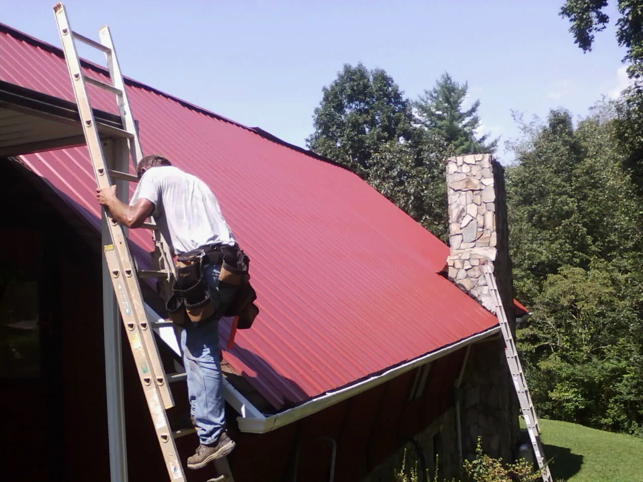 Roofer on a ladder beside a red metal roof and stone chimney — Soffit & Fascia Repair by Roofing Brothers in Borrego Springs